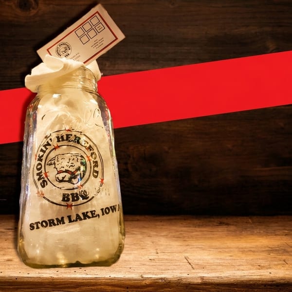 A Smokin' Hereford BBQ jar with a loyalty card on a wooden table with a red stripe.