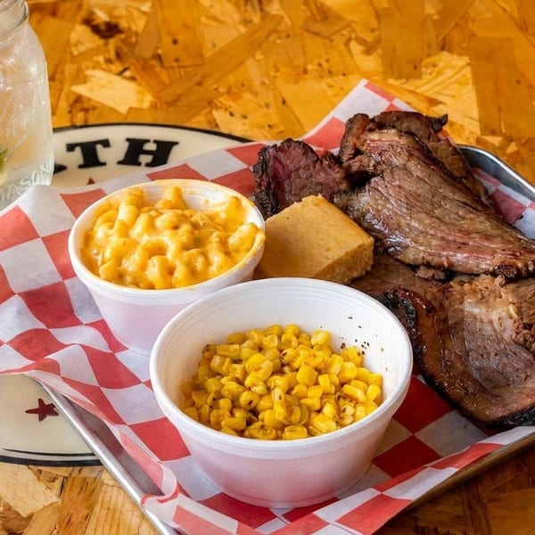 A BBQ platter with sliced brisket, mac and cheese, corn, cornbread, and a drink.