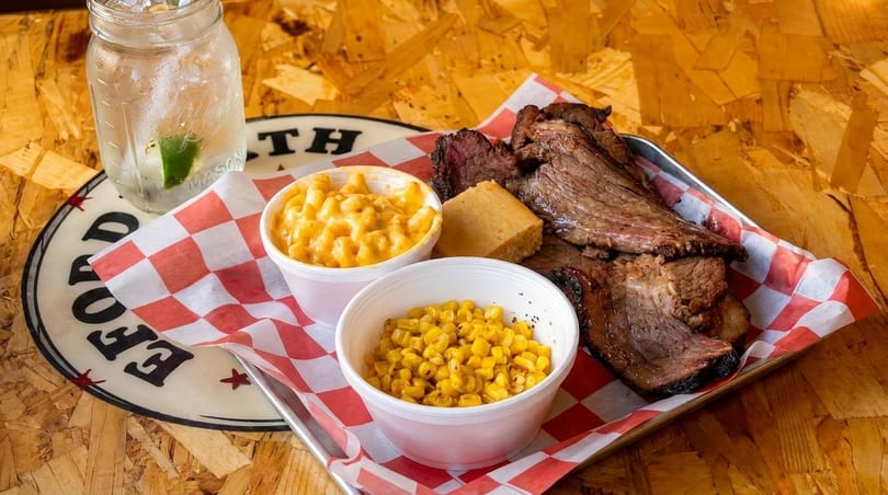 A BBQ platter with sliced brisket, mac and cheese, corn, cornbread, and a drink.