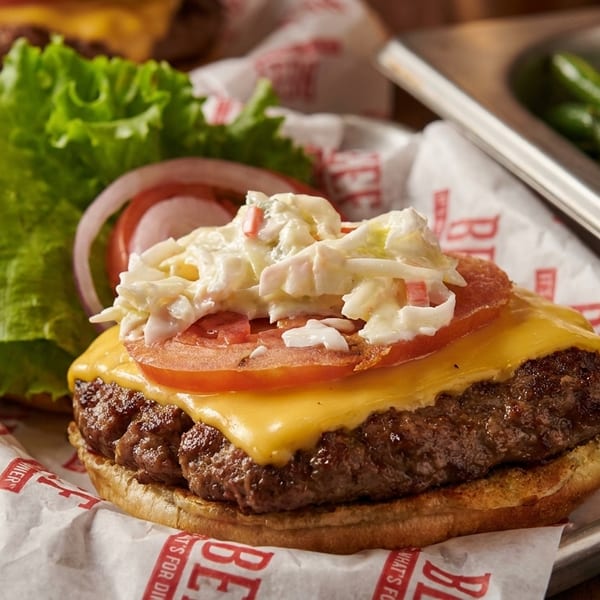 Cheeseburger with coleslaw, tomato, and onion on branded paper, with green beans in a metal tray.