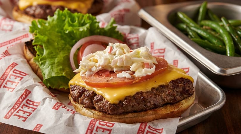 Cheeseburger with coleslaw, tomato, and onion on branded paper, with green beans in a metal tray.