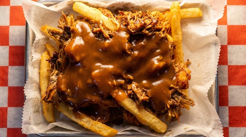 Close-up of golden french fries topped with pulled pork and rich brown gravy in a metal tray.