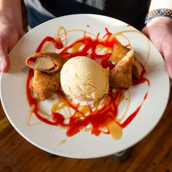 Hands holding a white plate with fried ice cream, strawberry sauce, and caramel drizzle.