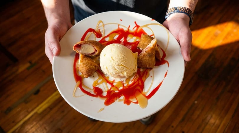 Hands holding a white plate with fried ice cream, strawberry sauce, and caramel drizzle.